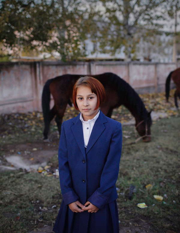 A girl with auburn colored brown hair wearing a blue school outfit with skirt, standing in front of a horse